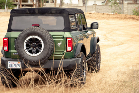 DV8 Offroad tube fender flares installed on green 4-door Ford Bronco, rear three-quarter view with spare tire near building
