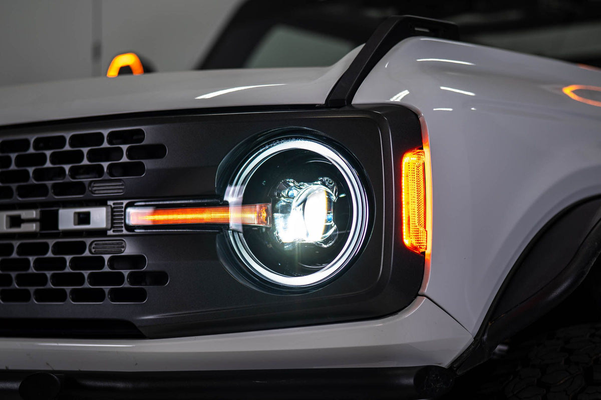 DV8 Offroad Black Series LED headlight installed on white Ford Bronco, angled close-up showing amber DRL bar and red marker light