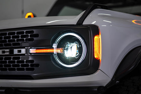DV8 Offroad Black Series LED headlight installed on white Ford Bronco, angled close-up showing amber DRL bar and red marker light