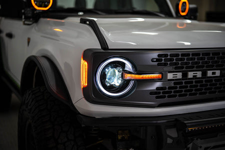 DV8 Offroad Black Series LED headlight installed on white Ford Bronco, close-up with DRL ring illuminated in shop setting