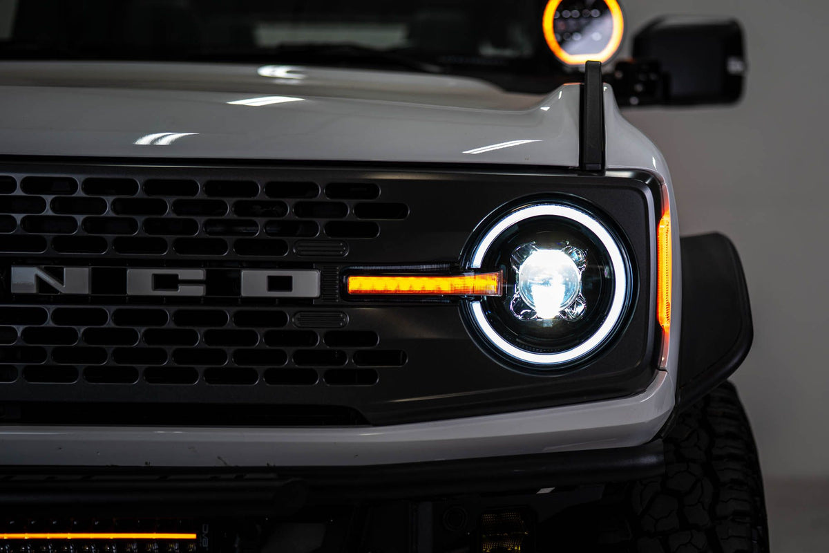 DV8 Offroad Black Series LED headlight installed on white Ford Bronco, close-up showing BRONCO grille badge and illuminated DRL ring