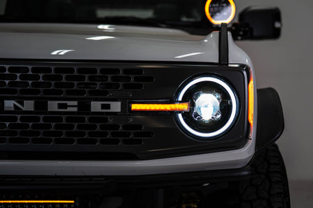 DV8 Offroad Black Series LED headlight installed on white Ford Bronco, close-up showing BRONCO grille badge and illuminated DRL ring