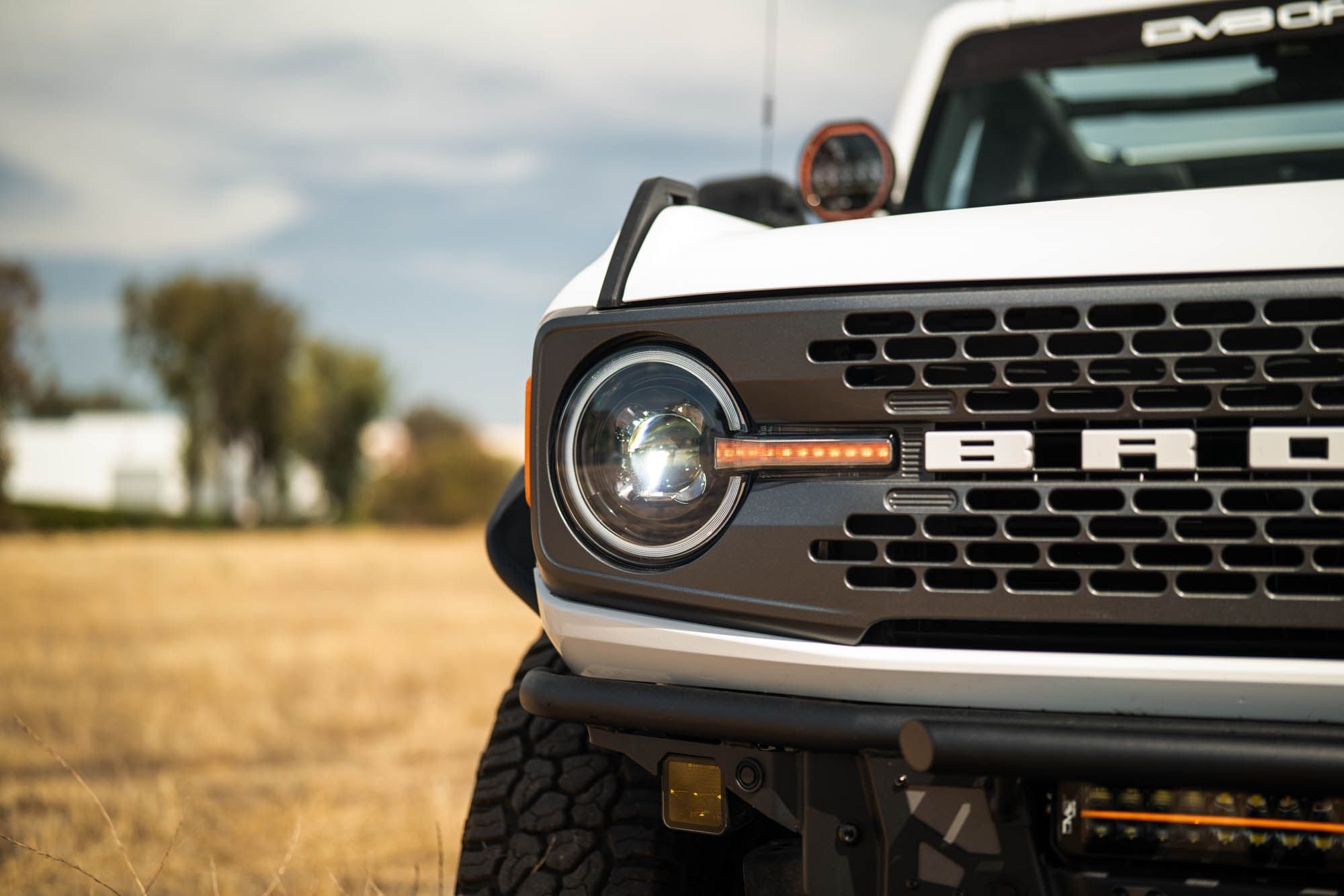 DV8 Offroad Black Series LED headlight installed on white Ford Bronco, close-up showing grille badge and headlight in field