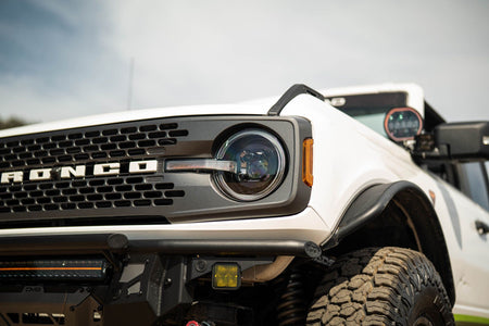 DV8 Offroad Black Series LED headlight installed on white Ford Bronco with DV8 Offroad windshield banner, front three-quarter close-up in field