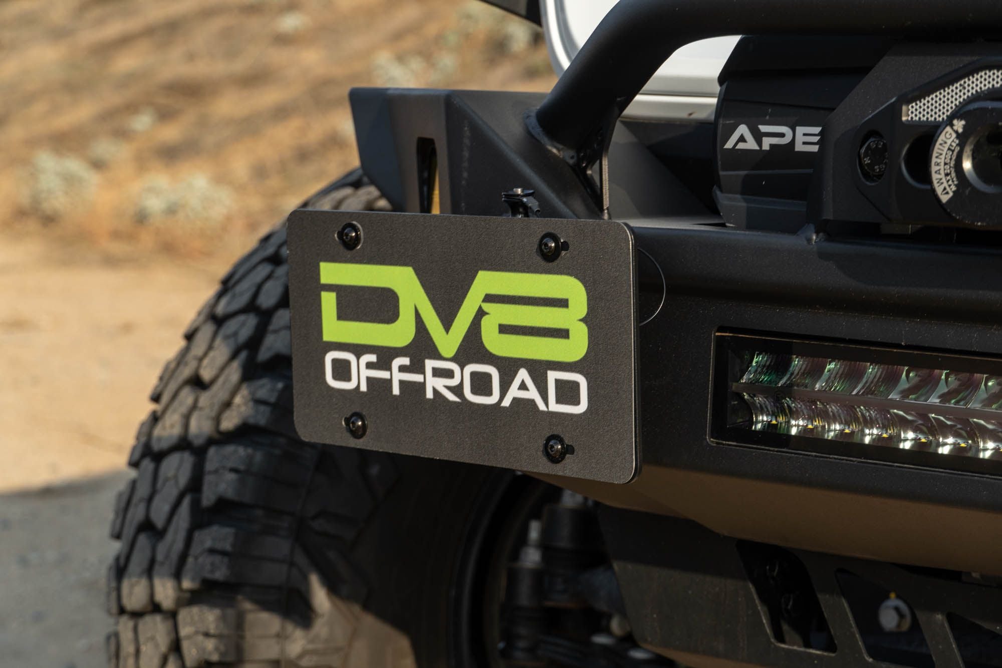 DV8 Offroad universal D-ring license plate mount installed on off-road bumper outdoor desert setting with DV8 light bar visible