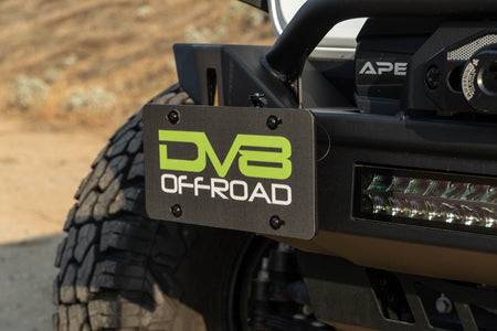DV8 Offroad universal D-ring license plate mount installed on off-road bumper outdoor desert setting with DV8 light bar visible