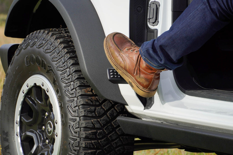DV8 Offroad foot peg in use on 2021-2026 Ford Bronco close-up showing foot stepping onto peg at door hinge mount