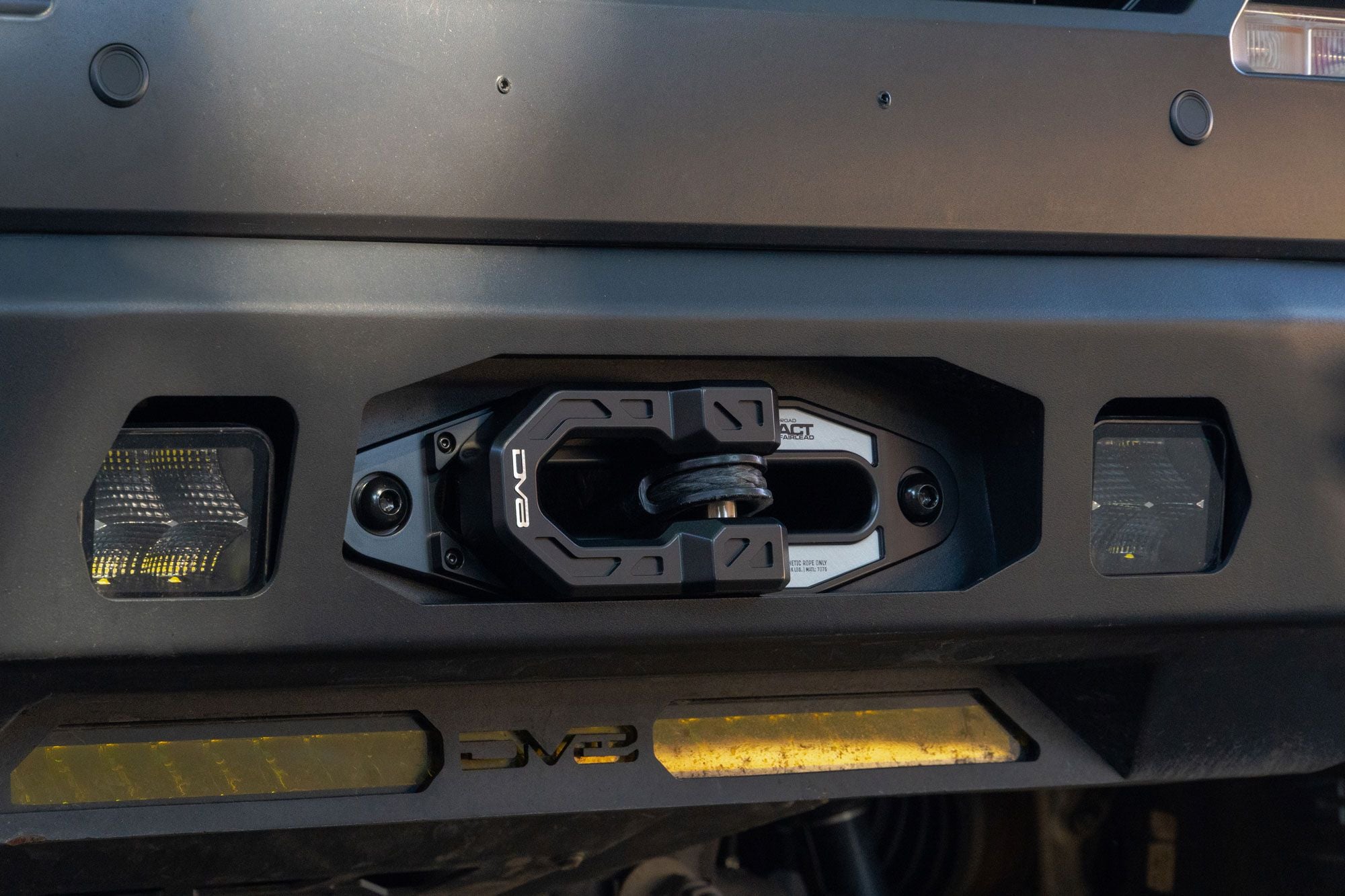 DV8 Offroad Xtract fairlead installed on dark gray truck front bumper, close-up with amber LED light bars and DV8 badge