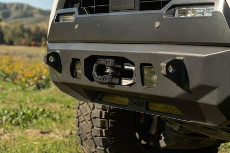 DV8 Offroad Xtract fairlead installed on olive green truck front bumper, wildflower field with mountains in background