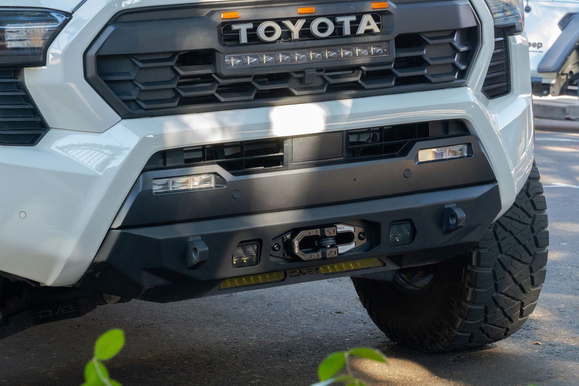 DV8 Offroad Xtract fairlead installed on white Toyota Tacoma front bumper, close-up front view with TOYOTA grille badge